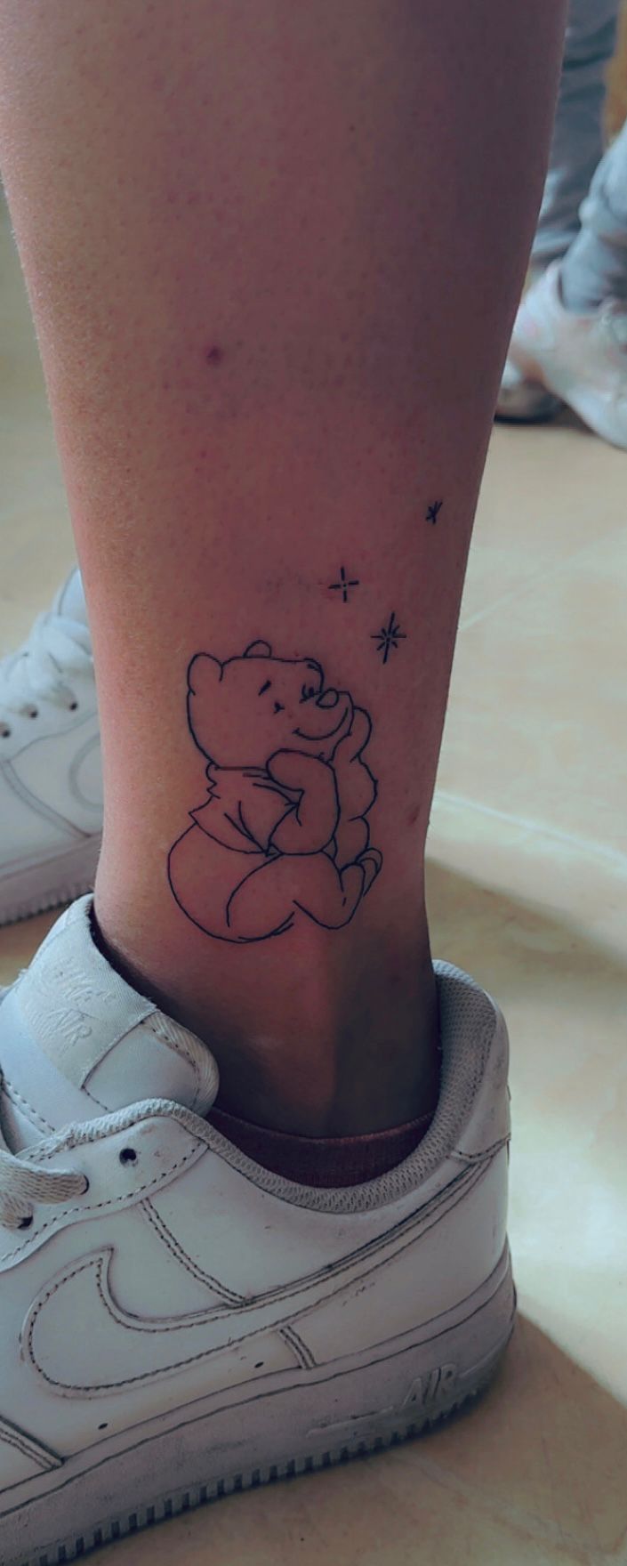Tatouage fineline Winnie l'Ourson assis avec etoiles sur la cheville Hakuna Tattoo Arpajon